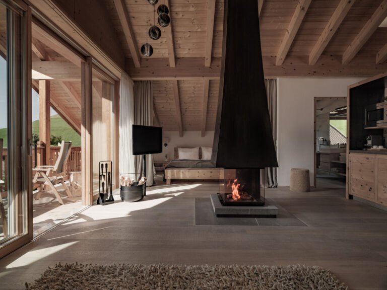 Open-plan natural-wood family suite at Sonnwies with a spruce double bed, a suspended black fireplace, a soft rug and sliding glass doors opening onto a balcony with green Alpine meadow views.