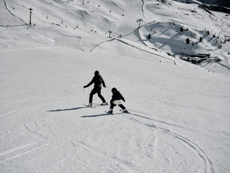 An adult skis alongside a small child on a wide, sunlit Dolomites piste with chairlifts and a lift station visible on the snowy slope behind them.