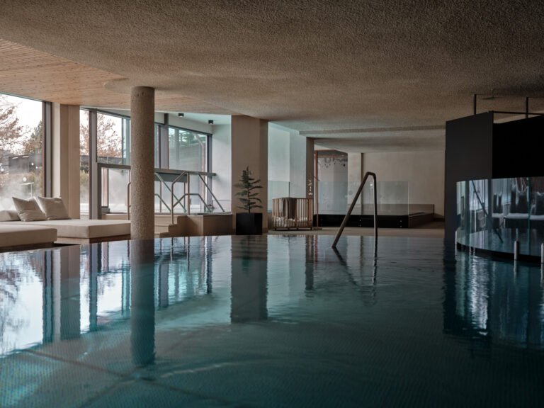 Steaming indoor pool at Sonnwies under a soft-formed ceiling, with floor-to-ceiling windows looking out to the surrounding mountains and a cushioned daybed at the water's edge.