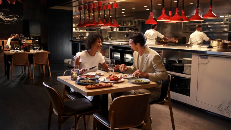 Couple dining at the chef's counter beside the open kitchen and red pendant lamps at Capa steakhouse