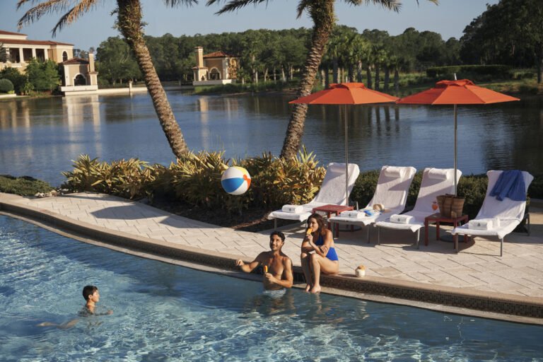 Family relaxing at the lakeside family pool with orange umbrellas and a beach ball, lake and palms behind