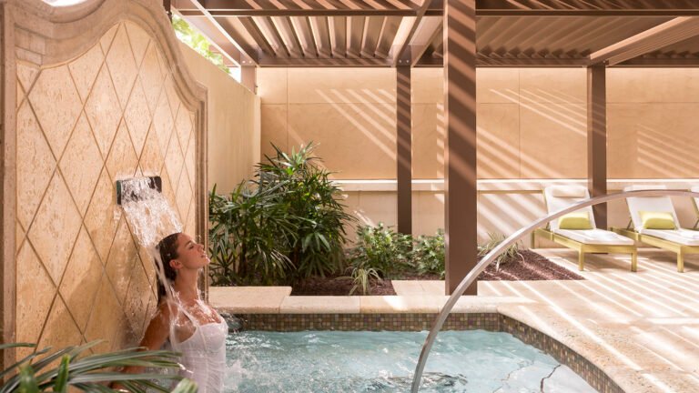 Guest under a waterfall rain shower in the spa's outdoor hydrotherapy garden with loungers