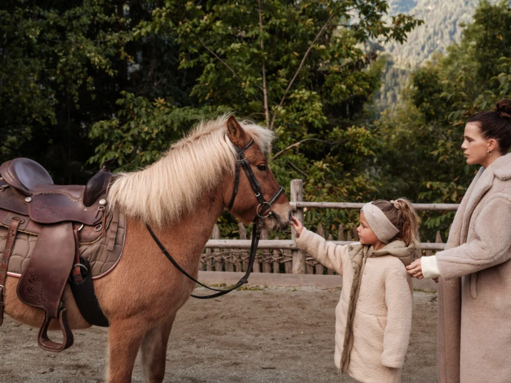 A young girl reaches out to stroke the nose of a Haflinger pony while her mother holds her hand, in a wooden paddock backed by the forested slopes of the Dolomites at Family Hotel Sonnwies in Luesen, South Tyrol.
