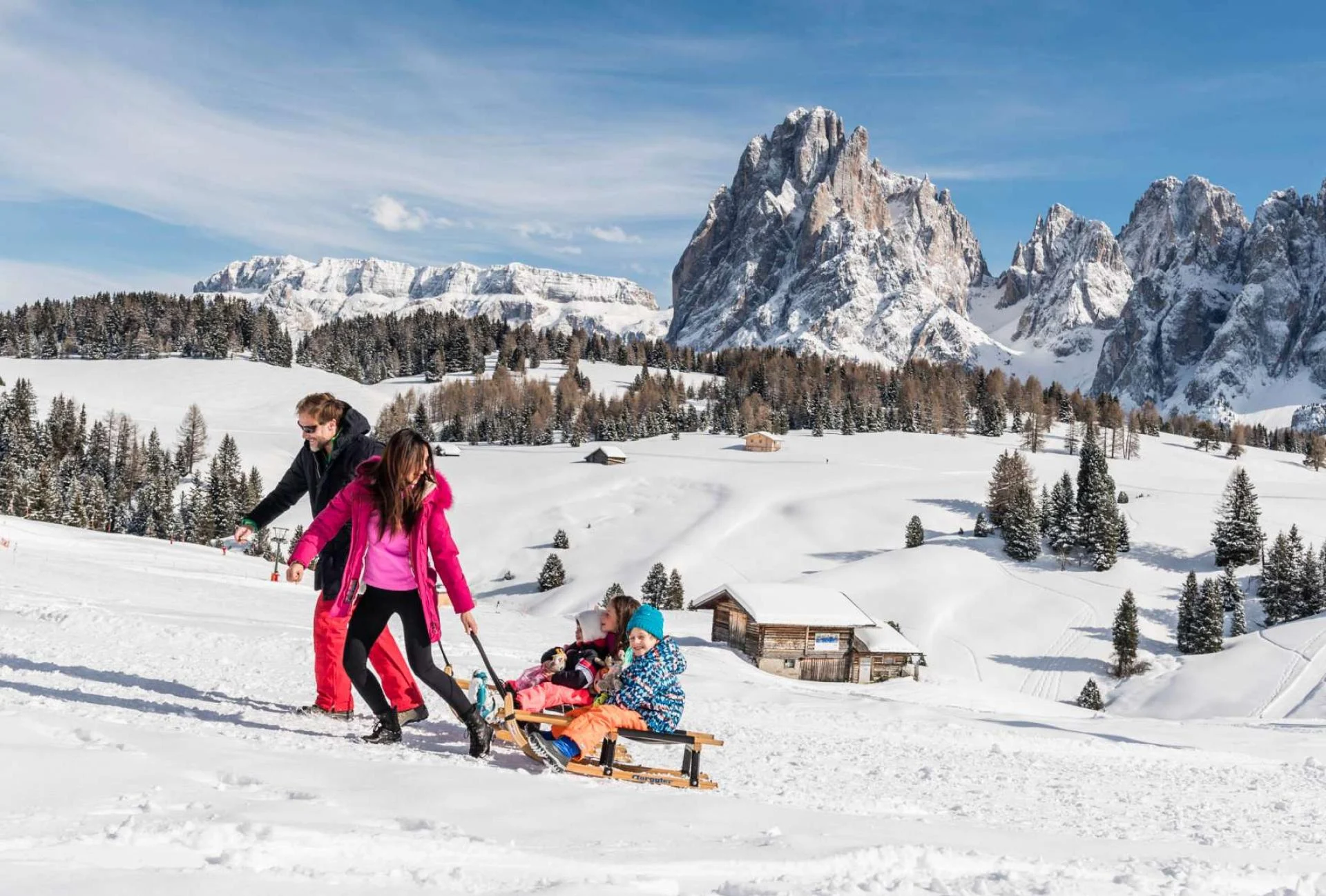 Family pulling two children on a sledge across snow with the Sassolungo Dolomites behind