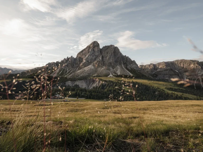 The jagged grey Dolomite peaks of the Puez-Geisler massif reflected in a still alpine lake at golden hour, the valley floor leading toward the Sonnwies estate far below.