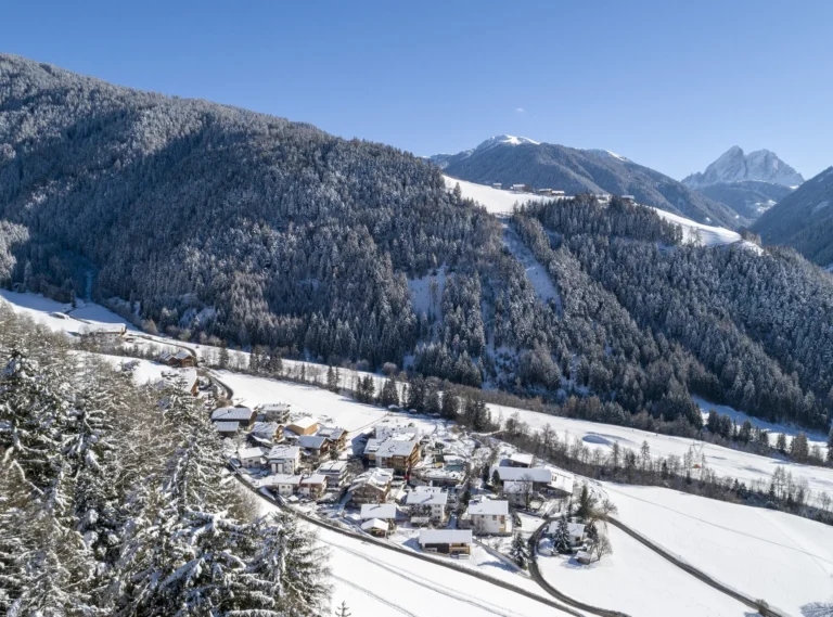 A drone aerial photograph of the Family Hotel Sonnwies complex from above, the stone-and-timber buildings and outdoor pool set on a hillside terrace surrounded by pine forest, with the Lüsen valley and Dolomite peaks stretching to the horizon.