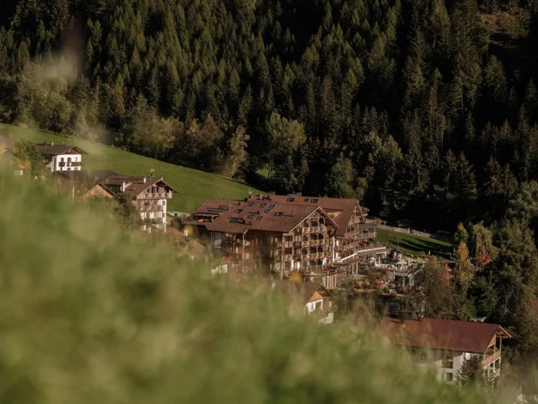 A wide panoramic shot of the Lüsen valley at dawn, the Sonnwies hotel building nestled on a terraced hillside with meadows below and the Dolomite rock faces glowing pink in the early light.