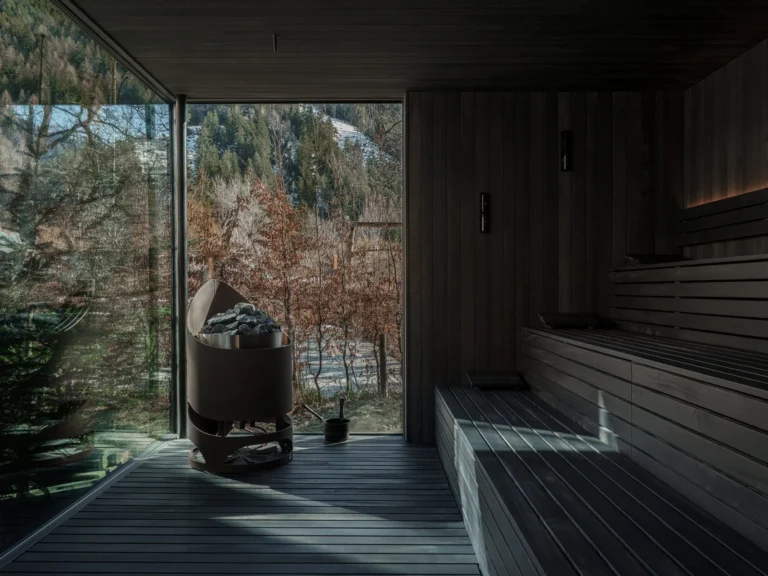 A wellness treatment room at Sonnwies with a wide window overlooking the mountain forest, a therapist preparing a stone-pine oil beside a cushioned treatment table.