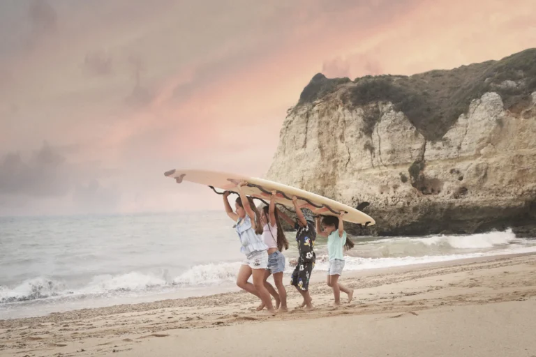 Kinder tragen gemeinsam ein Surfboard am Strand Praia dos Tremoços