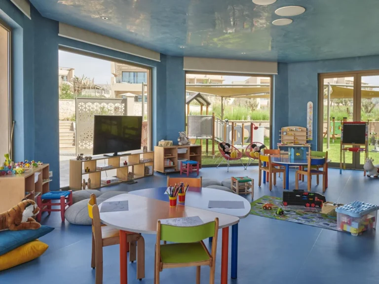 Bright indoor kids' club room with colourful low tables, chairs, toy storage and floor-to-ceiling windows overlooking the grounds