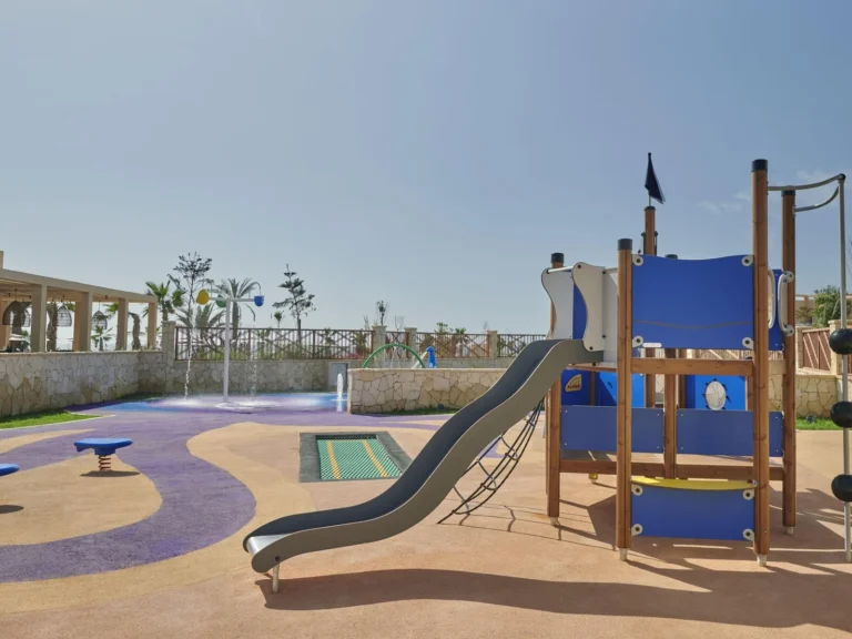 A pirate-ship-themed outdoor playground with a slide, climbing rope and spring rockers on a purple-and-sand-colored splash-safe surface