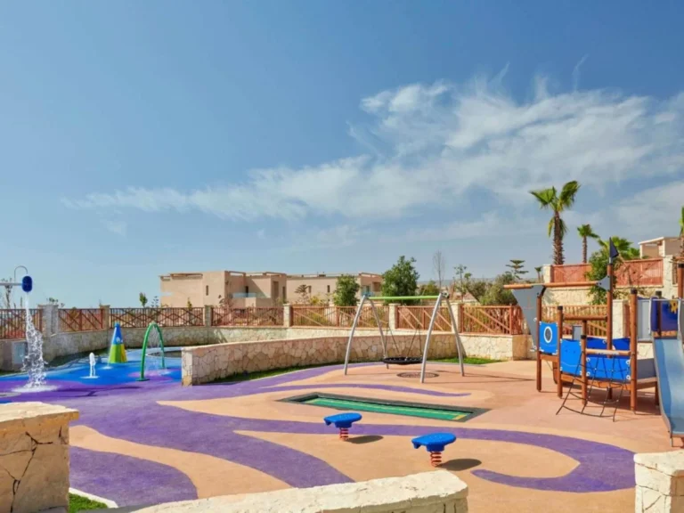 A splash pad with water jets next to a swing set in the outdoor kids' play area, framed by palm trees