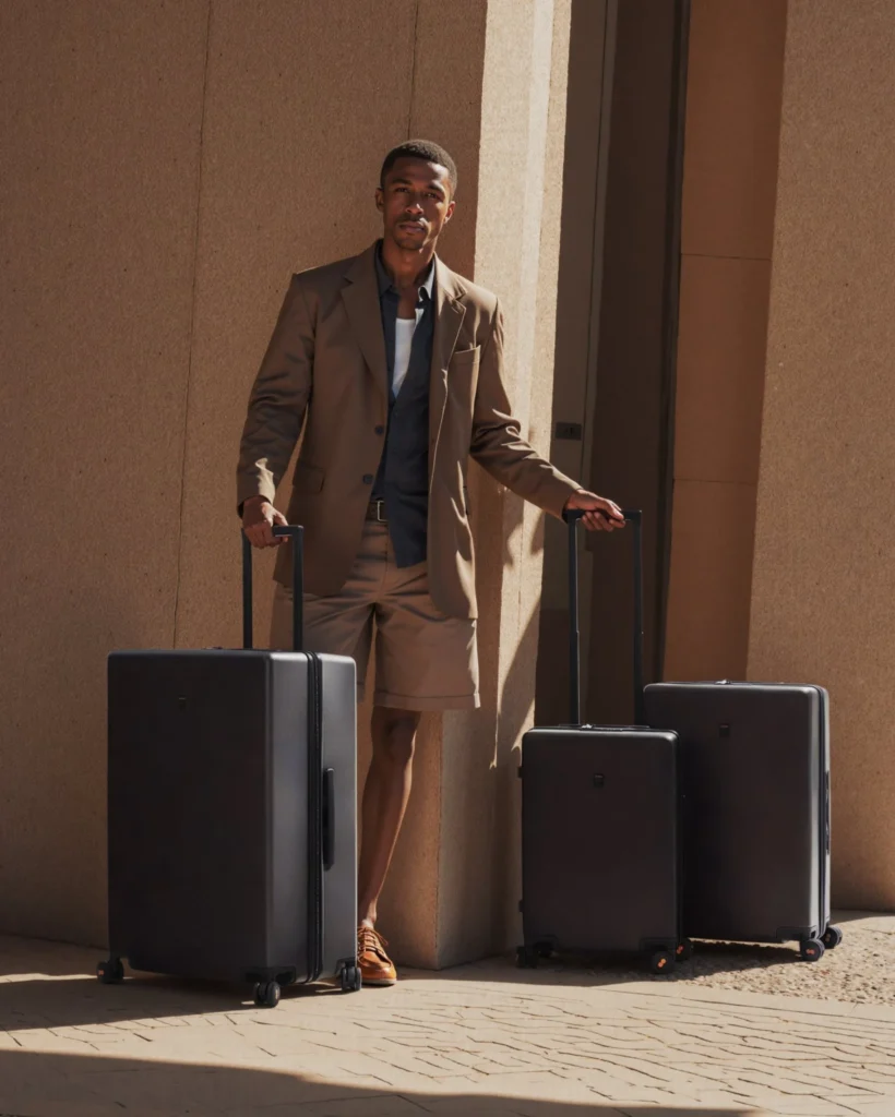 A traveller stands between two LEVEL8 Luminous Textured hardshell suitcases, telescopic handles raised, against a sunlit stone facade