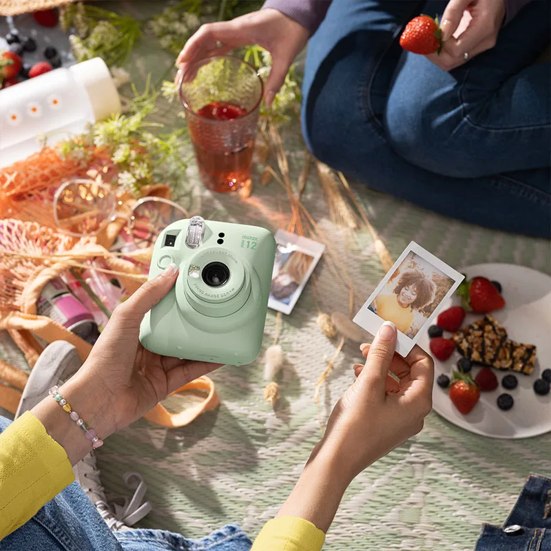 Two people at a picnic holding a Mint Green Fujifilm Instax Mini 12, one showing a freshly printed instax photo