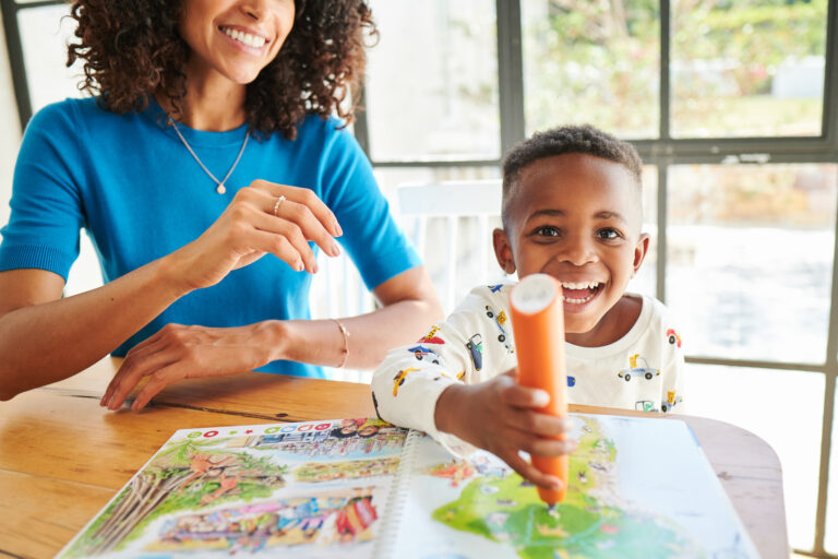 Young boy laughing with joy while using tiptoi pen on an open tiptoi book, mother watching beside him at kitchen table