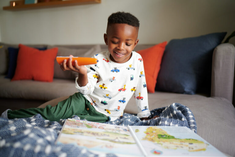 Young boy sitting cross-legged on sofa holding tiptoi pen over an open tiptoi book, smiling with delight