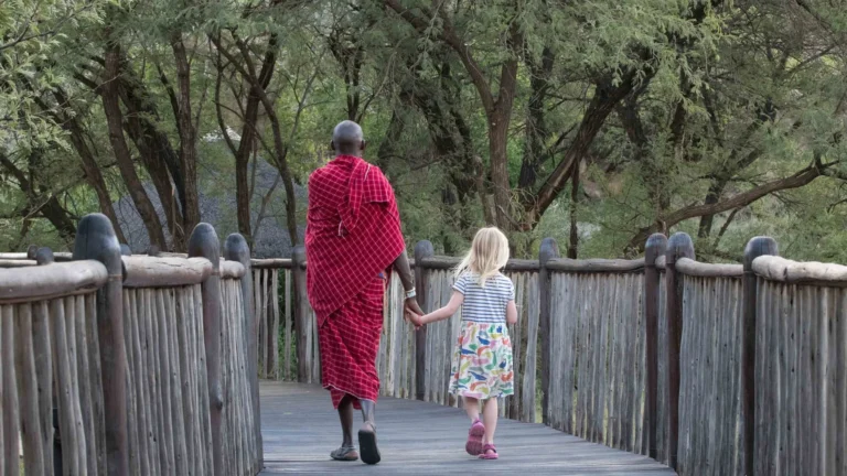 Young guest and Maasai host crossing a raised walkway through the lodge grounds