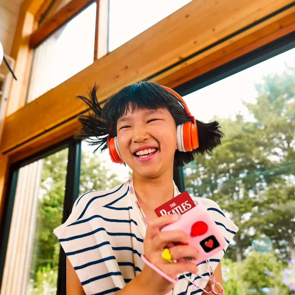 Smiling child holding a Yoto Mini in a pink Adventure Jacket while wearing orange headphones