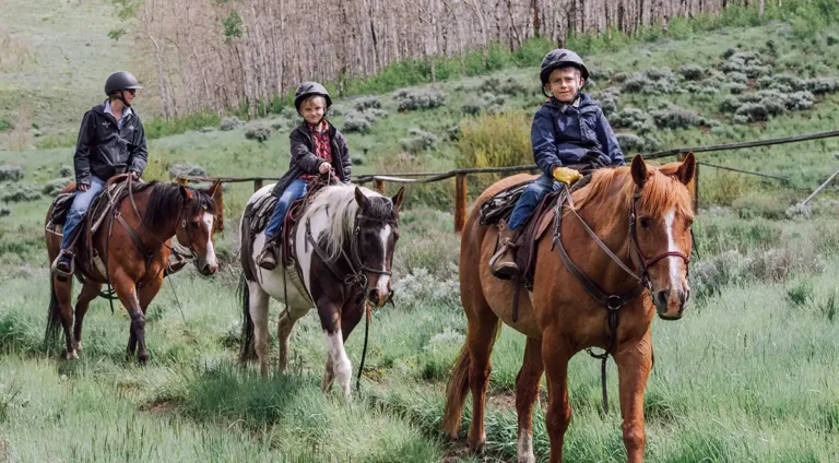 Three children in riding helmets on horseback along a wooden fence line at C Lazy U Ranch