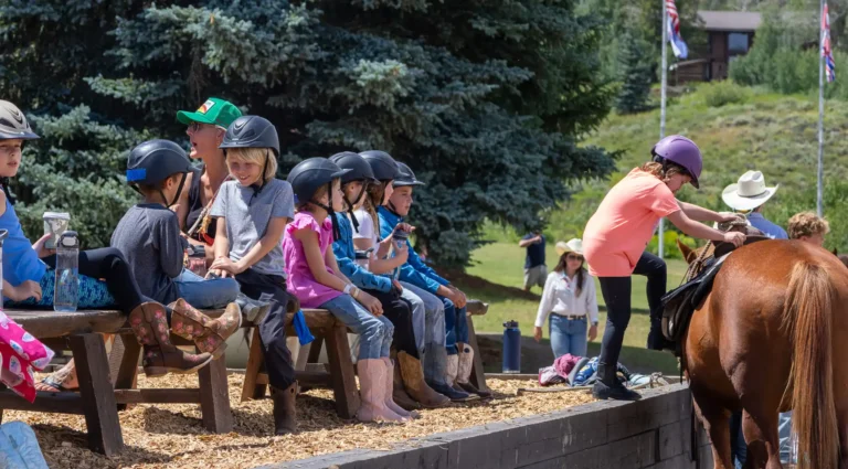 Children in riding helmets line up at the mounting block, assisted by wranglers, before a trail ride