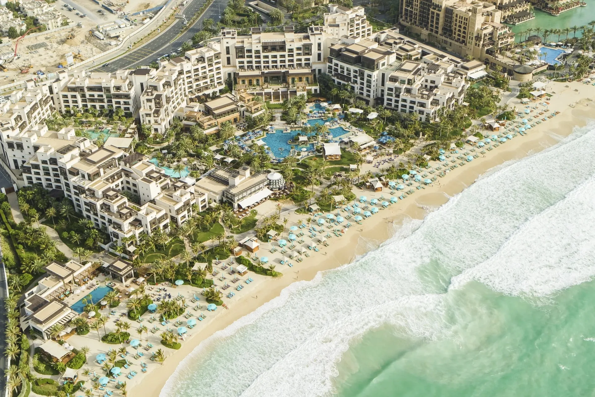 Wide aerial view of the full Jumeirah Al Naseem resort complex — white Arabic-influenced buildings, multiple free-form pools, lush tropical gardens and the beachfront all visible from above