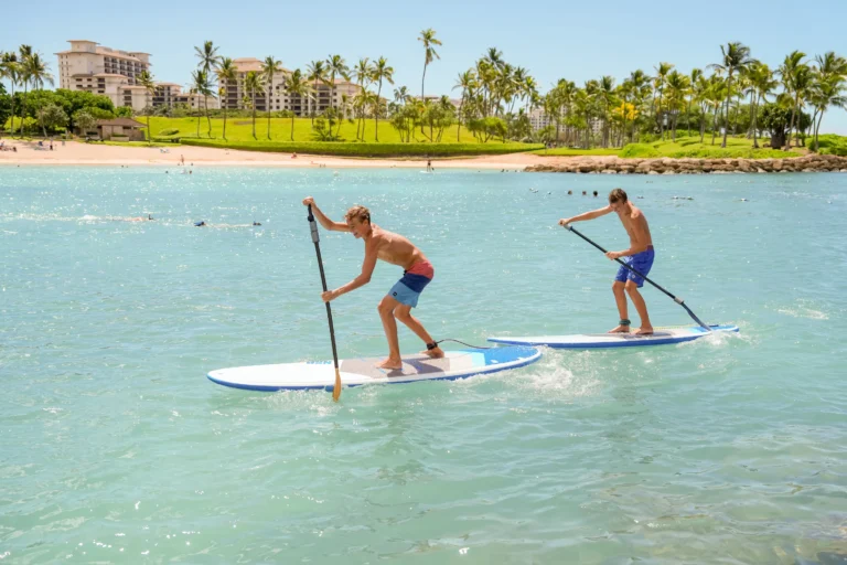 Two teenage boys stand-up paddleboard together in the turquoise waters of Ko Olina lagoon with the resort building behind
