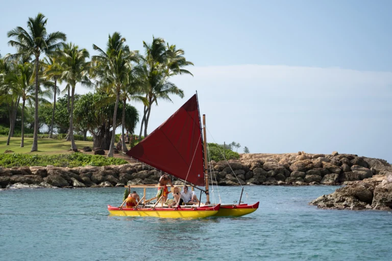 Traditional Hawaiian outrigger sailing canoe with red sail on Ko Olina lagoon with the resort grounds, palms and rocks behind