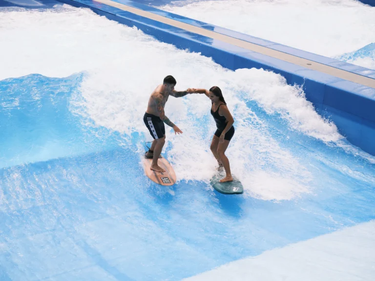Couple surfing side by side on the FlowRider wave simulator at Ko Olina, holding hands on the artificial wave