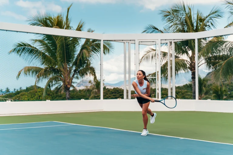 Woman plays tennis on the blue hard court at Four Seasons Resort Oahu with palm trees and Ko Olina mountains behind