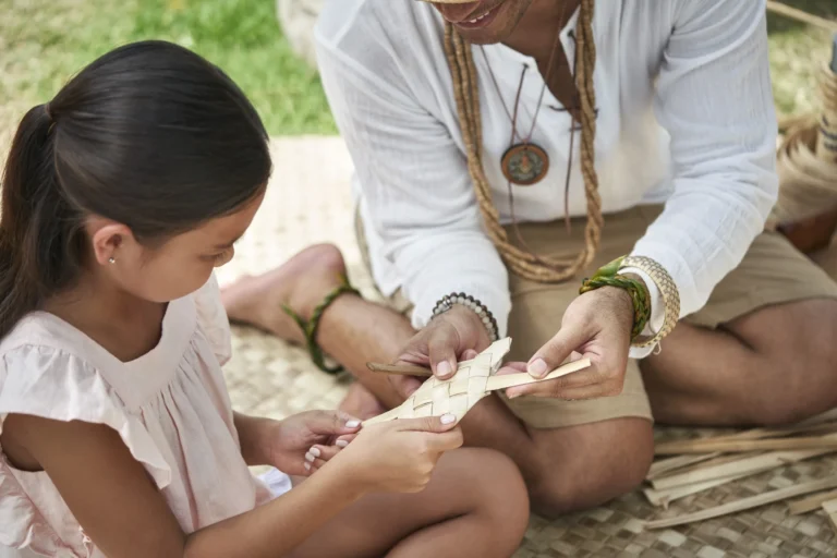 Young girl learns traditional Hawaiian lauhala palm weaving from a cultural practitioner at Camp Kohola, Four Seasons Resort Oahu