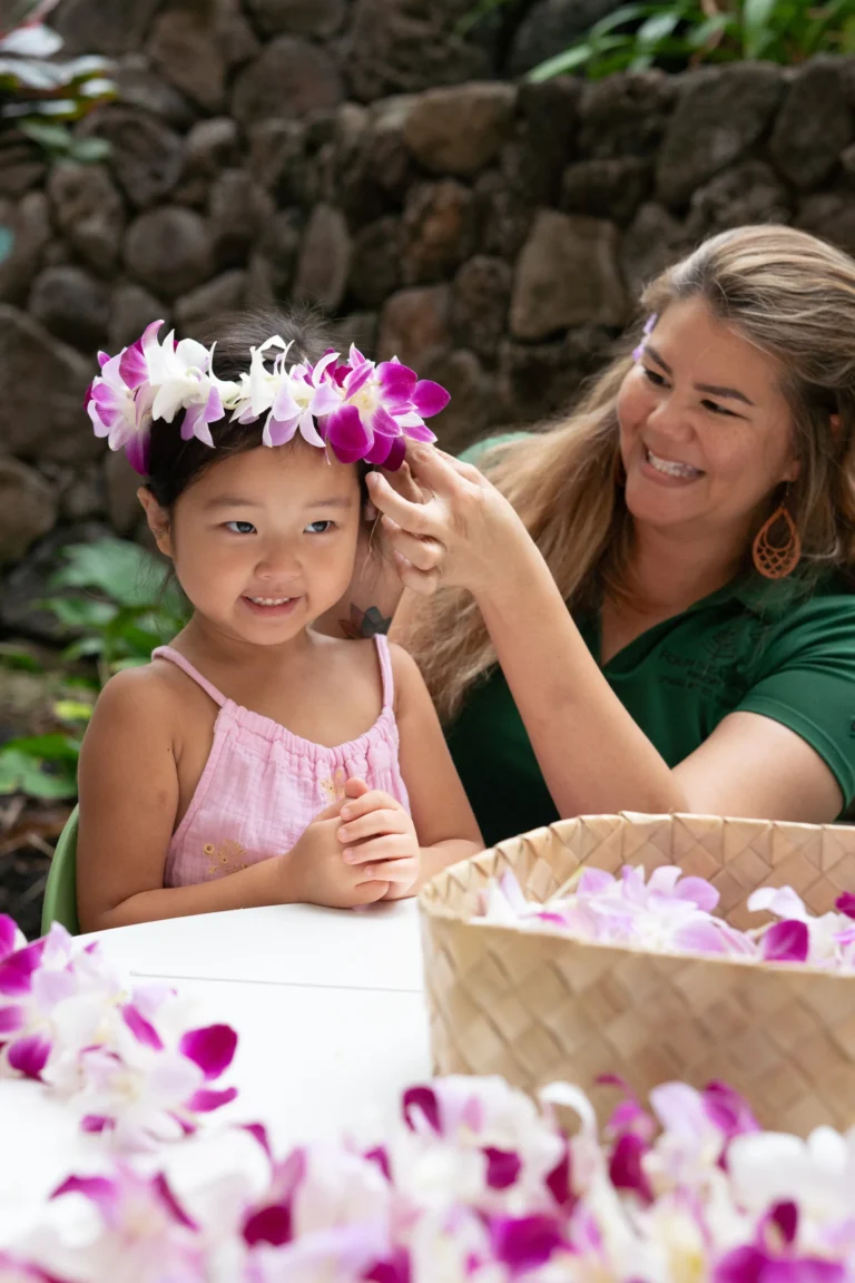 Resort cultural staff places a fresh flower crown on a smiling young girl during a lei and flower-crown making session