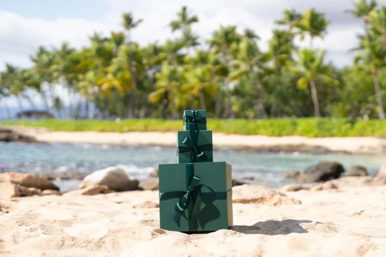 Three stacked green holiday gift boxes on Ko Olina beach sand with the lagoon and palm trees behind
