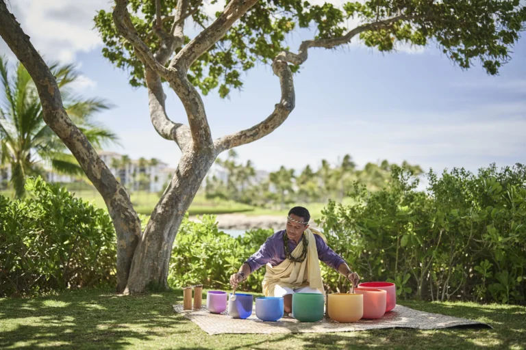 Hawaiian cultural wellness practitioner plays colourful crystal singing bowls on the resort grounds beneath a large tree at Ko Olina