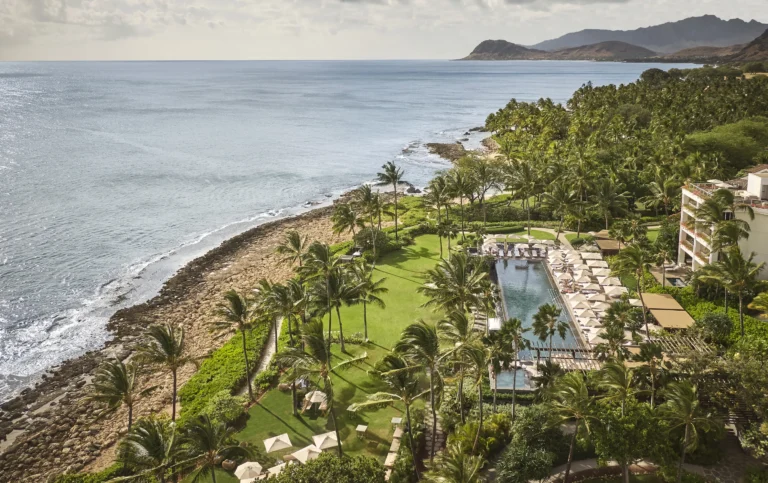 Aerial view of Four Seasons Resort Oahu infinity pool, palm-lined lawn and leeward coastline stretching toward the Waianae mountains
