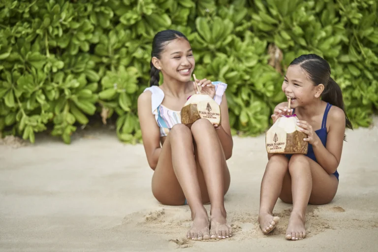 Two girls sitting on the sand drinking from fresh coconuts at Four Seasons Resort Oahu