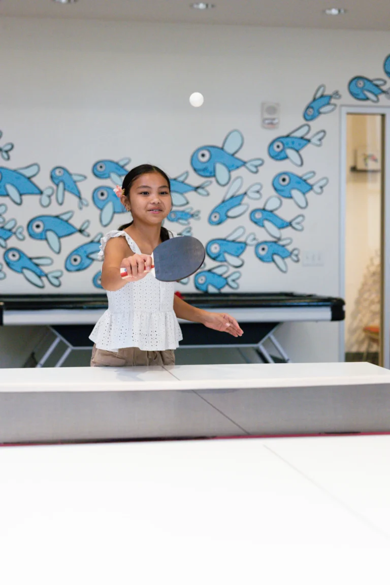 A girl playing table tennis in the Camp Kohola games room with its painted fish mural