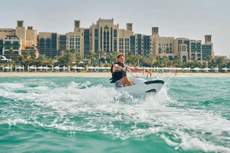 Teenage boy on a jet ski in the Arabian Gulf, grinning as he rides through turquoise water, with the Madinat Jumeirah Arabic windtower buildings and the hotel's white beach parasols visible behind him on the shore