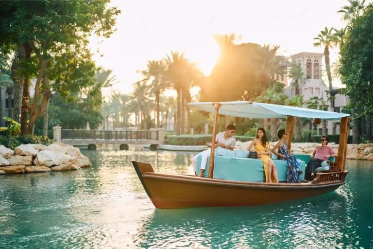 Four adult guests seated on a traditional wooden abra boat with mint-green upholstered cushions gliding along the Madinat Jumeirah waterway at golden hour, palm trees and Arabic windtower buildings lining the banks behind them