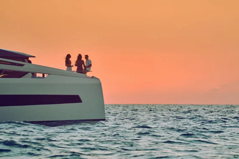 Three guests silhouetted against a vivid deep-orange sunset sky, standing on the bow deck of a luxury white motor yacht in the Arabian Gulf, champagne glasses in hand