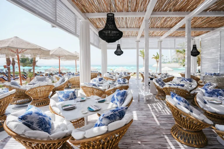 Open-air Summersalt Beach Club restaurant at Jumeirah Al Naseem with rattan pod chairs, navy-and-white tropical-print cushions, a thatched ceiling with black pendant chandeliers and direct views to the Arabian Gulf beach