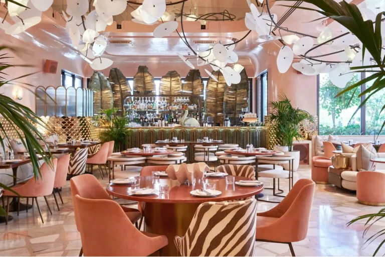 Flamingo-pink dining room at Jumeirah Al Naseem with blush velvet chairs, zebra-print accents, a dramatic sculptural white floral ceiling installation with black metalwork branches, and a full bar fronted by giant golden banana-leaf panels