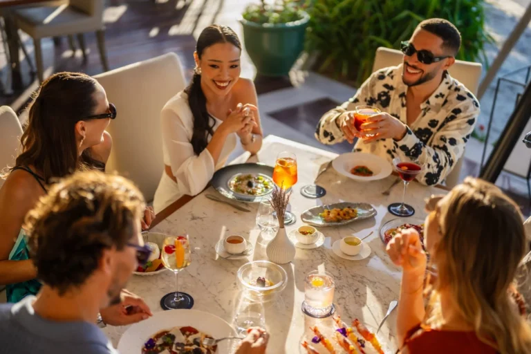 Five friends laughing and toasting over Aperol spritzes and colourful cocktails at a sunlit marble outdoor restaurant table at Jumeirah Al Naseem, with sharing plates of seafood between them