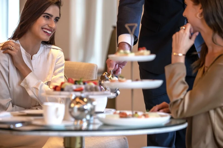 Two women smiling as a white-gloved waiter presents a silver three-tiered stand of petit fours, macarons and pastries at an elegantly set afternoon tea table inside Jumeirah Al Naseem