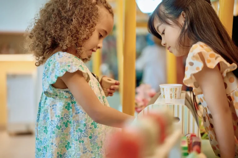 Two young girls aged approximately three and five, in floral and polka-dot dresses, focused and smiling as they play with a wooden toy tea set at a colourful activity station inside Sinbad's Kids Club at Jumeirah Al Naseem