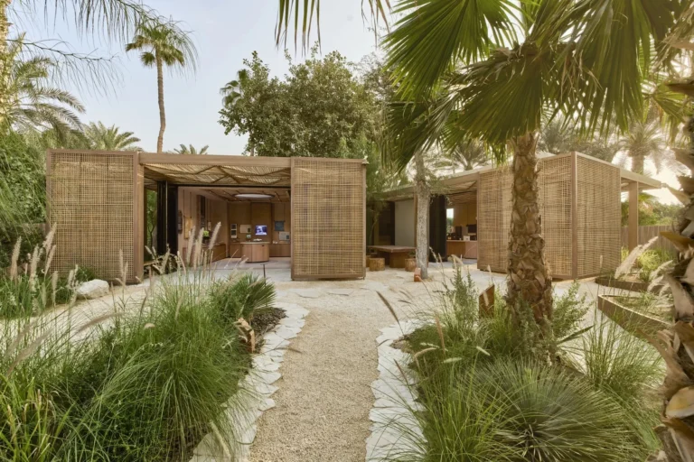 Exterior of Sinbad's Kids Club eco pavilion at Jumeirah Al Naseem — open bamboo-screen structures set among lush date palms, frangipani trees and ornamental grasses along a sandy stone path
