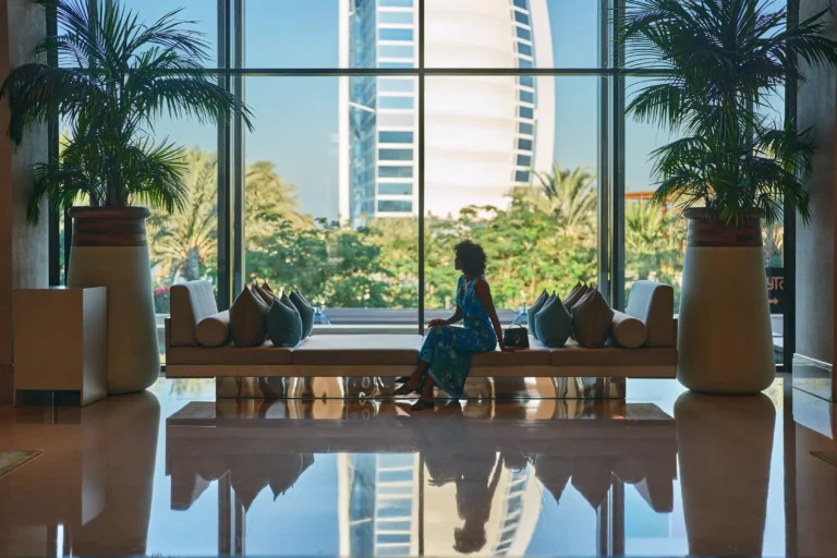 Jumeirah Al Naseem lobby with mirror-polished marble floors, large tropical palms in ceramic urns, and a woman in a blue dress seated on a marble bench silhouetted perfectly against full-height glass windows framing the Burj Al Arab
