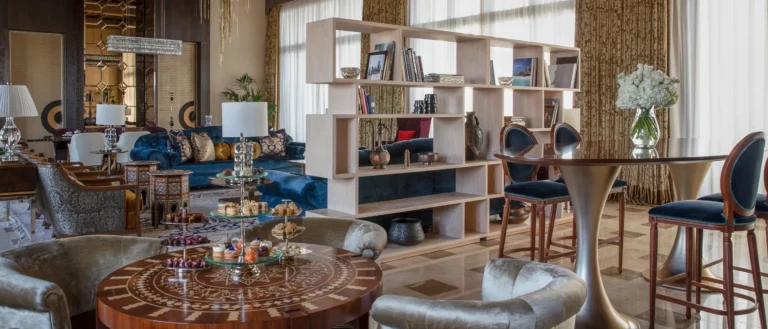 Ornate Arabian-style hotel lounge with blue velvet sofas, a tiered silver stand of pastries on an inlaid Moorish table, crystal chandeliers, and a bookshelf partition dividing the seating areas
