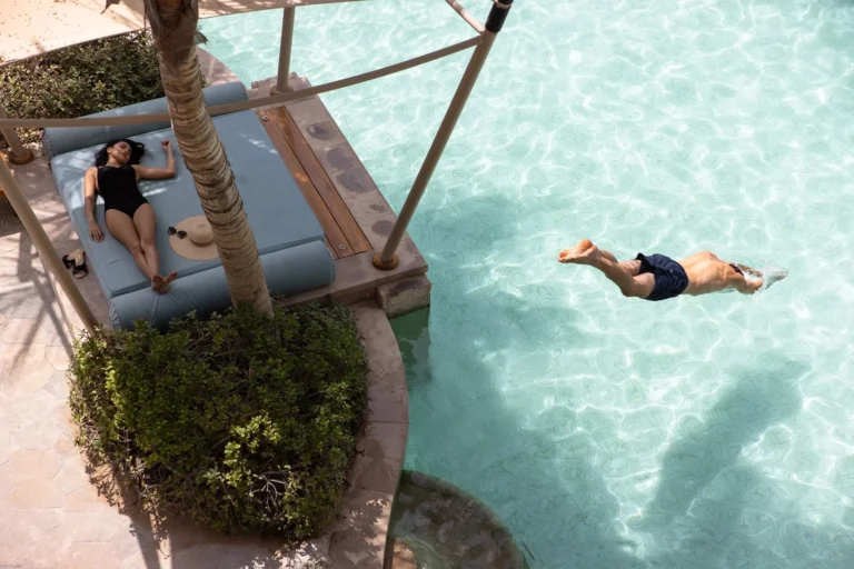 Overhead view of Jumeirah Al Naseem's lagoon pool — a woman sunbathing on a circular island platform daybed beside a palm tree while a man dives into the crystalline turquoise water