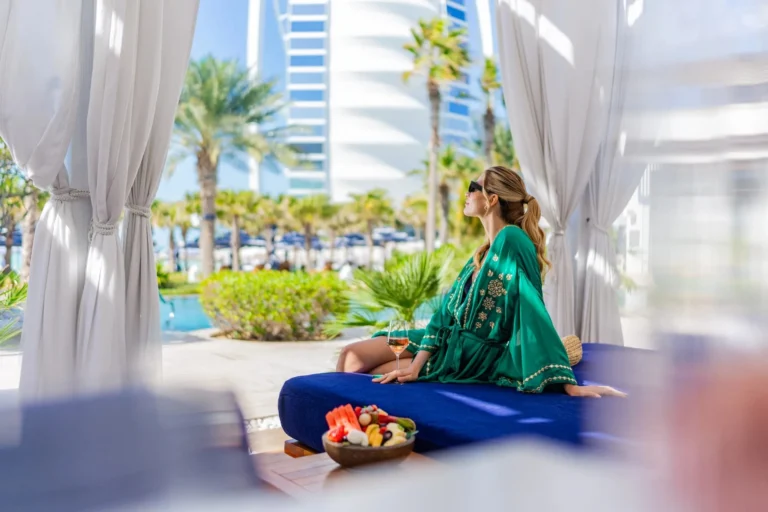 Woman in a green embroidered kaftan relaxing on a white upholstered daybed in a private pool cabana at Jumeirah Al Naseem, holding a wine glass with fruit on the table, the Burj Al Arab towering in the background above the palm-lined pool