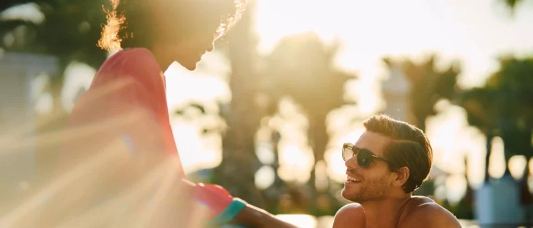 Couple relaxing in a Jumeirah Al Naseem pool at golden hour, palm trees silhouetted against a warm sunlit sky, the turquoise water glowing in the late-afternoon light